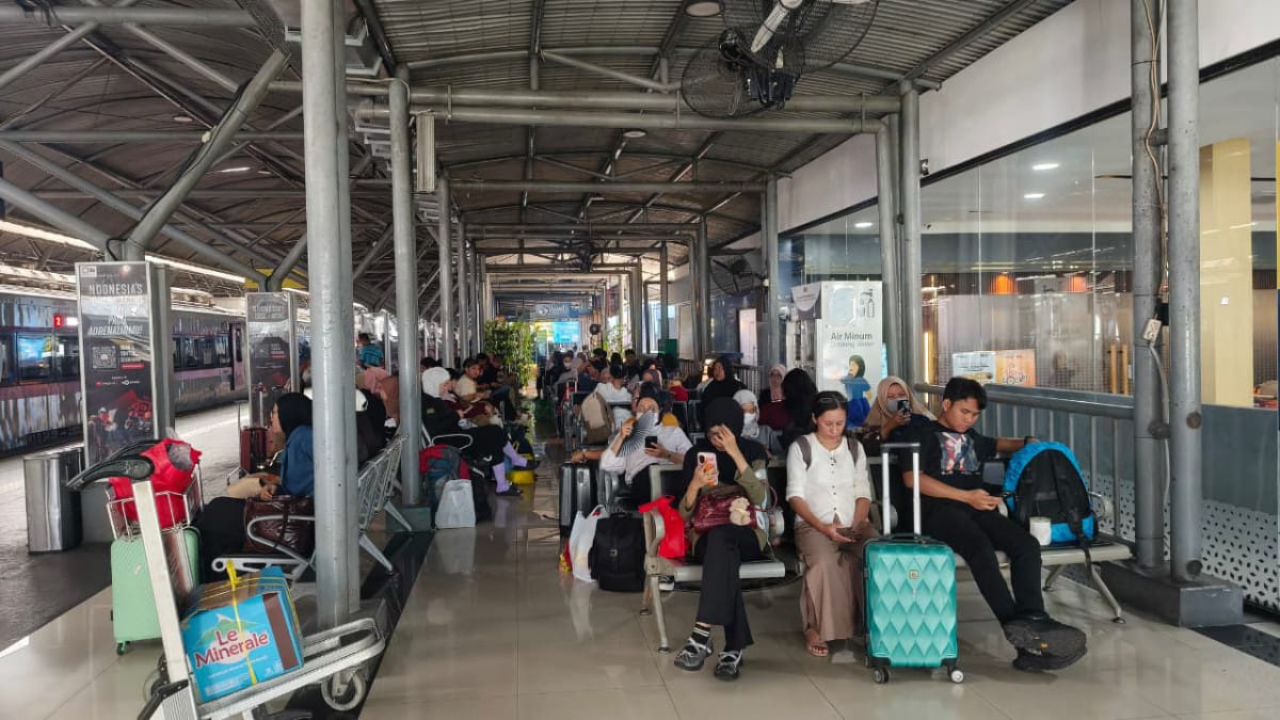 Penumpang kereta api menunggu keberangkatan di area stasiun dengan membawa barang bawaan dan koper.